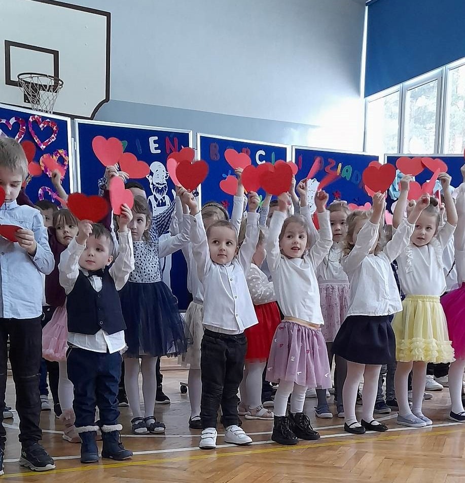 Przedszkolaki na niebieskim tle trzymające czerwone serca z papieru wysoko nad głowami.