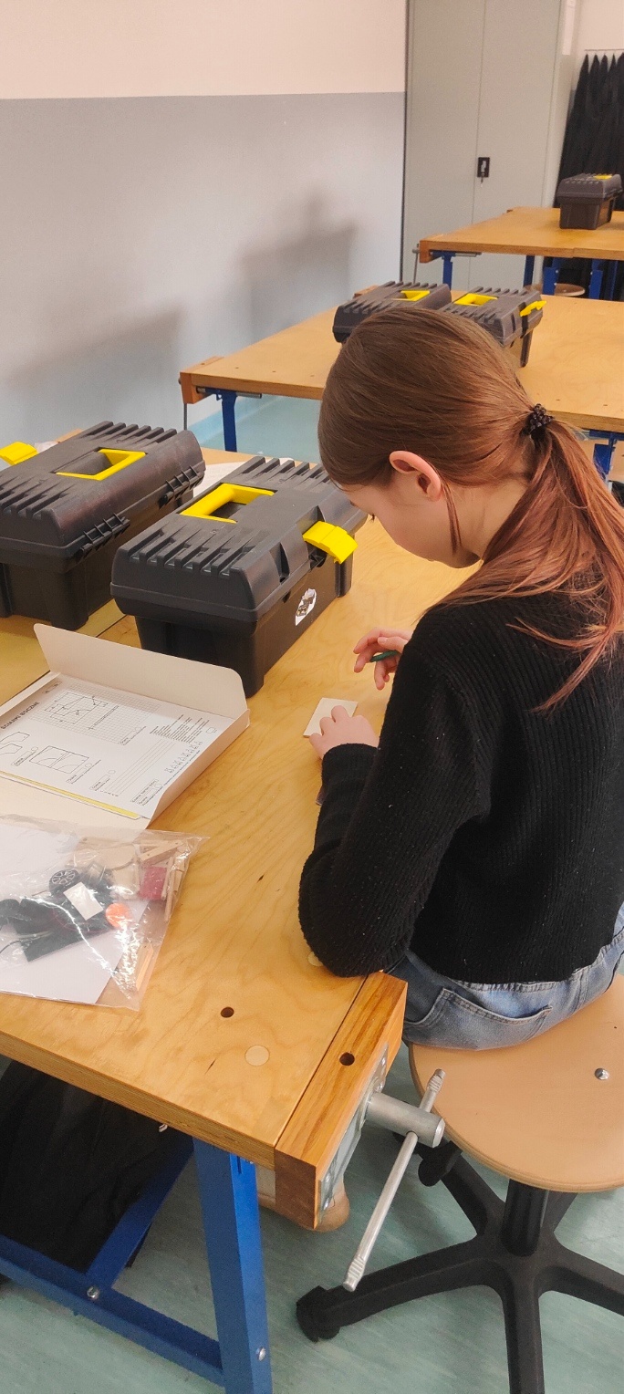Uczennica siedzi bokiem przy dużym drewnianym stole w szkolnej pracowni technicznej. Jest odwrócona tyłem i pochylona nad małym elementem, który trzyma w dłoniach. Na stole stoją czarne skrzynki narzędziowe z żółtymi uchwytami, leży też otwarte pudełko z kartą pracy i woreczek z drobnymi materiałami. W tle widać kolejne stoły i jasnoszarą ścianę.