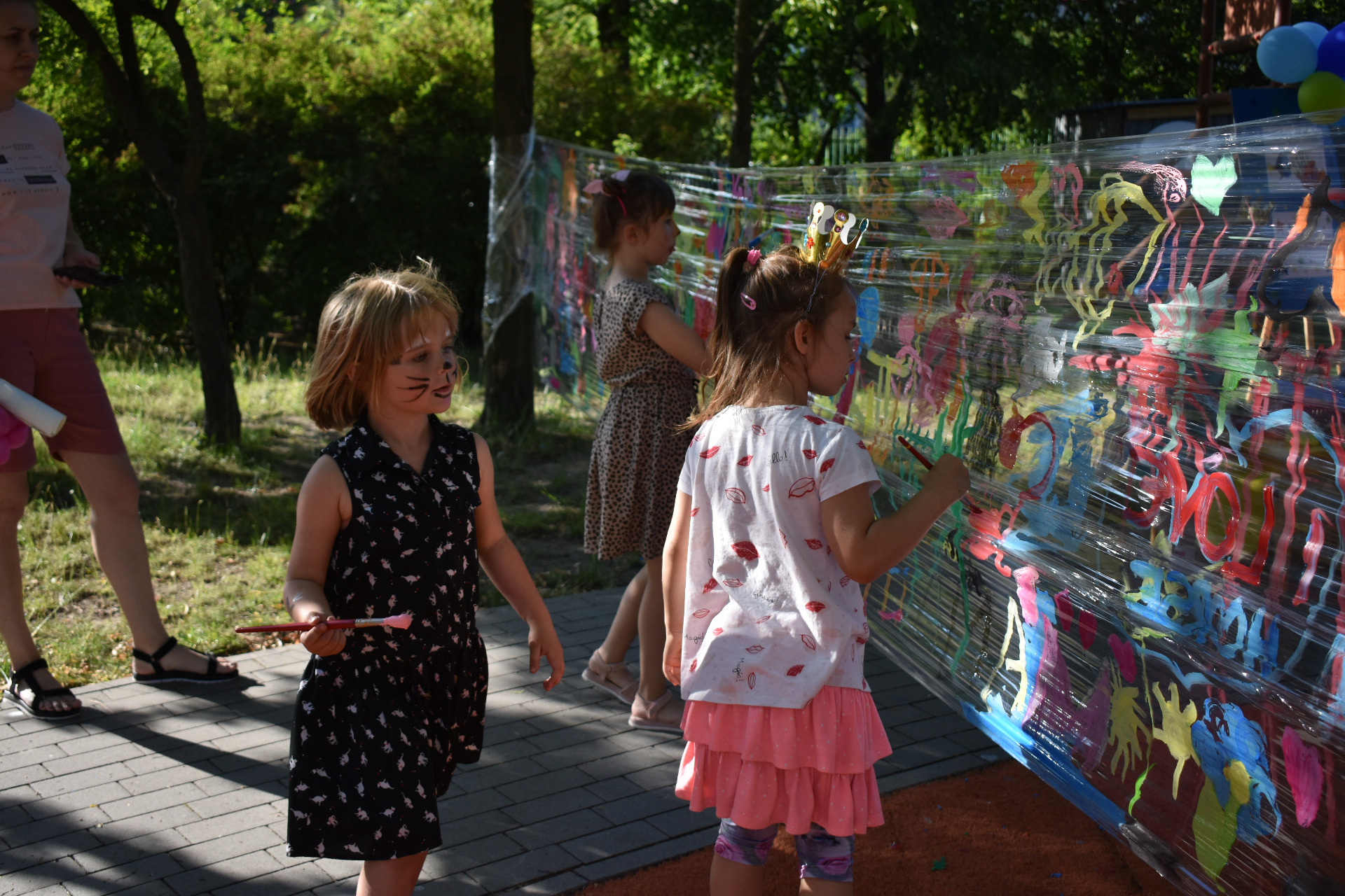Trzy dziewczynki malują farbami na przeźroczystej folii rozciągniętej między drzewami.