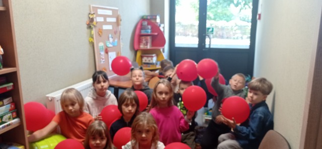 12 uczniów z balonikami siedzi na dywanie.