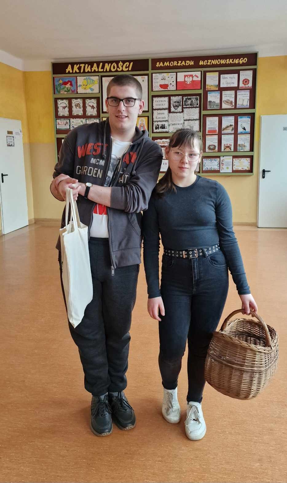 Uśmiechnięta nastolatka oraz nastolatek (oboje w okularach)  pozują z koszykiem i zwykłą torbą na zakupy