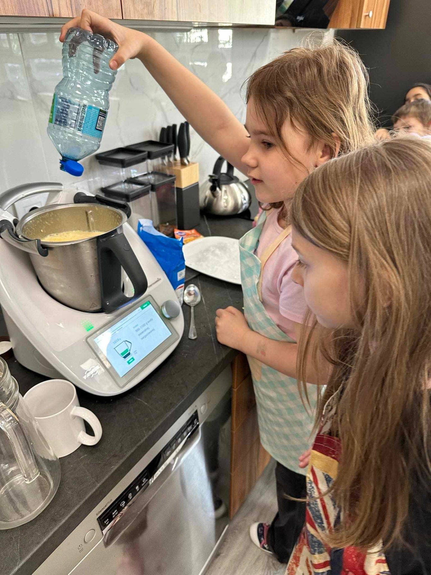 Na zdjęciu widzimy scenę w kuchni, gdzie dwójka dzieci zaangażowana jest w działania kulinarno-edukacyjne. Skupiamy się na dziecku, które nalewa wodę z przezroczystej plastikowej butelki.