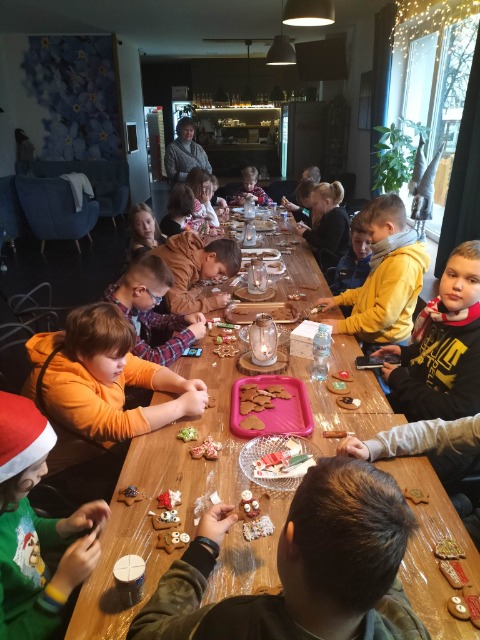 16 uczniów siedzi przy stole, jeden ma na głowie czapkę  Świętego Mikołaja. Na stole leżą pierniki i tace z piernikami.