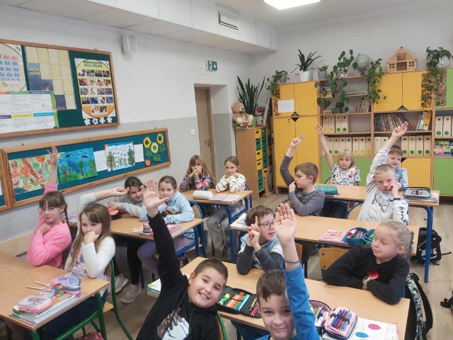 Uczniowie siedzą w sali w ławkach, część dzieci trzyma podniesioną do góry rękę, na stolikach leżą przybory szkolne, podręczniki.