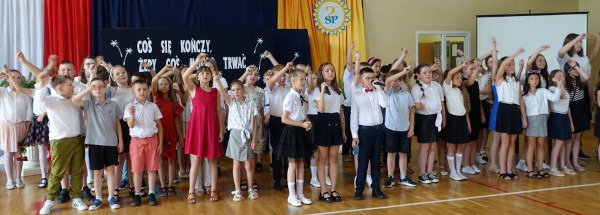 Duża grupa uczniów klas trzecich odświętnie ubranych podczas apelu stoi na sali gimnastycznej