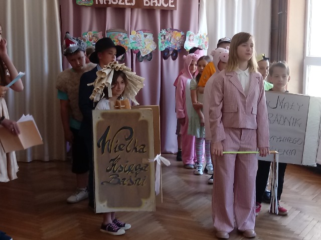 Z przodu stoją dwie dziewczynki, jedna z nich trzyma różdżkę, a druga karton z napisem: Wielka Księga Baśni. Dziewczynka ma na głowie kapelusz zrobiony ze zwiniętych kartek papieru. Za nimi w dwóch rzędach stoją dzieci.

