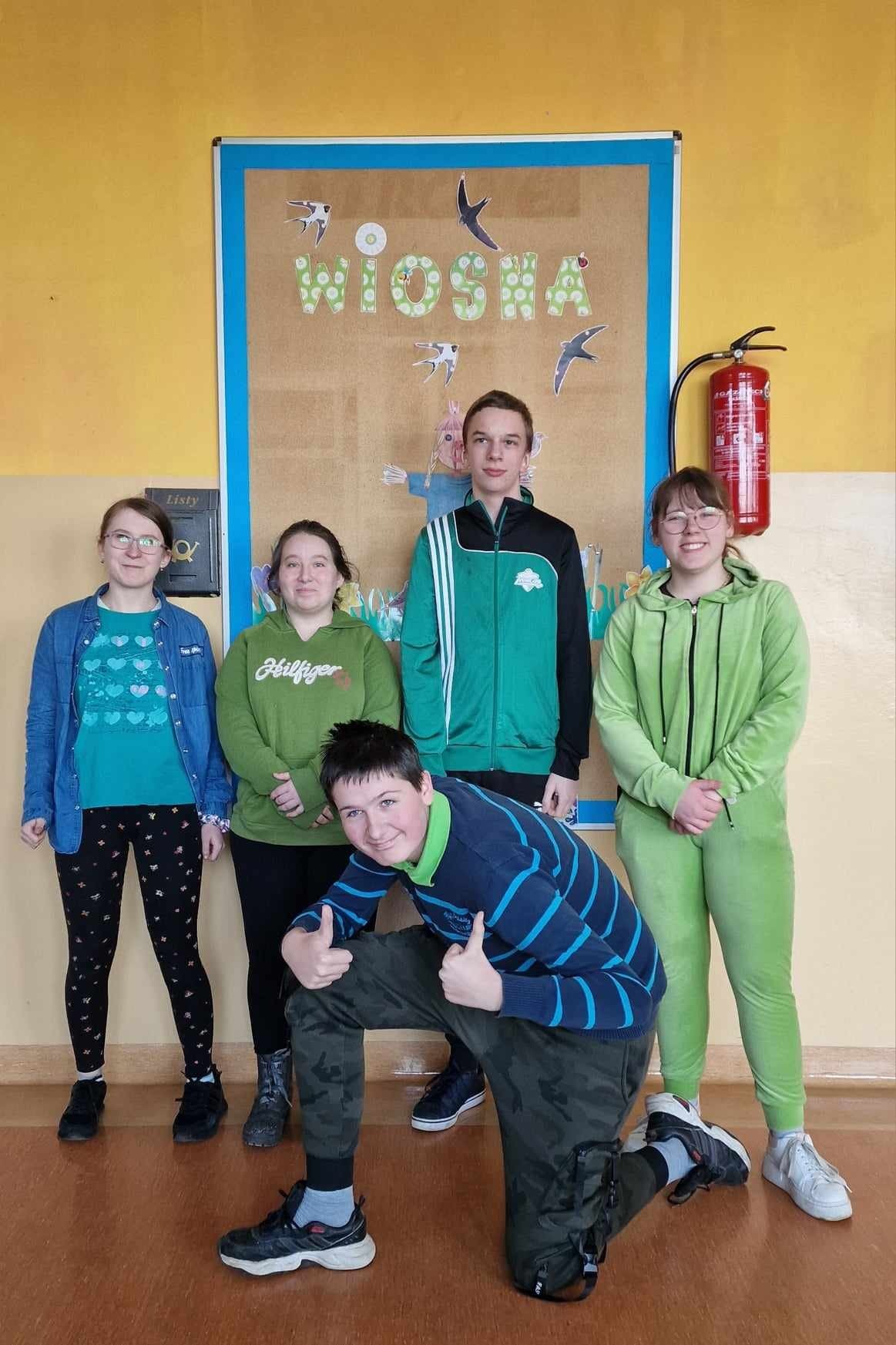 5 uczniów nastolatków ubranych na zielono pozuje przed gazetką ścienną pt. "Wiosna"